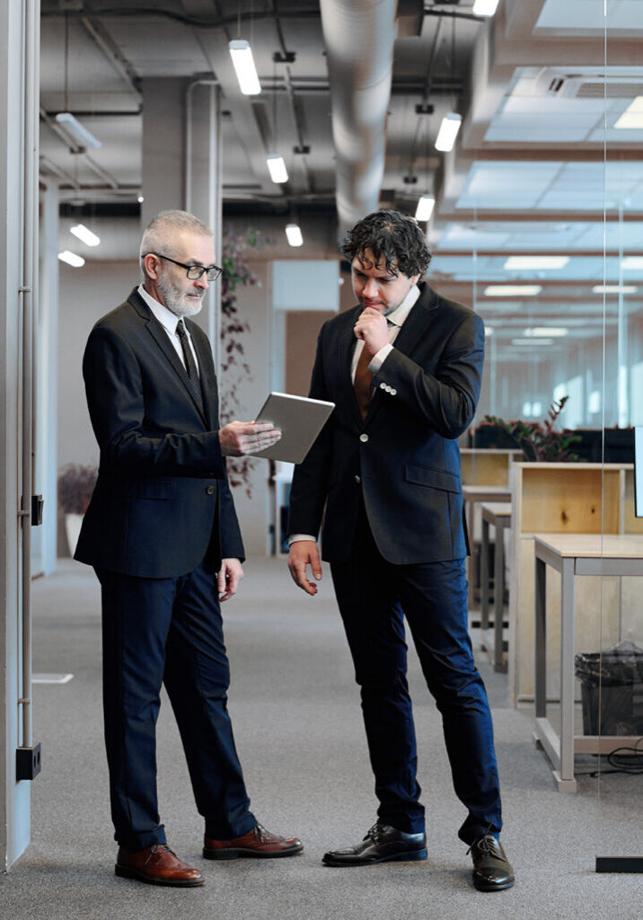 Mature businessman in suit showing something on digital tablet to his colleague while they standing at office